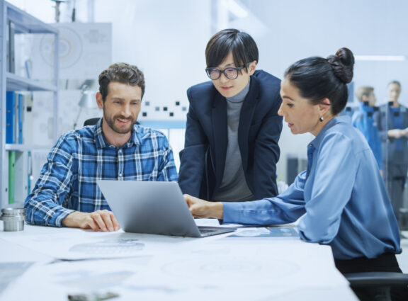 RFA Engineering team members collaborating on a project, gathered around a laptop to discuss technical plans and designs in a modern office environment.