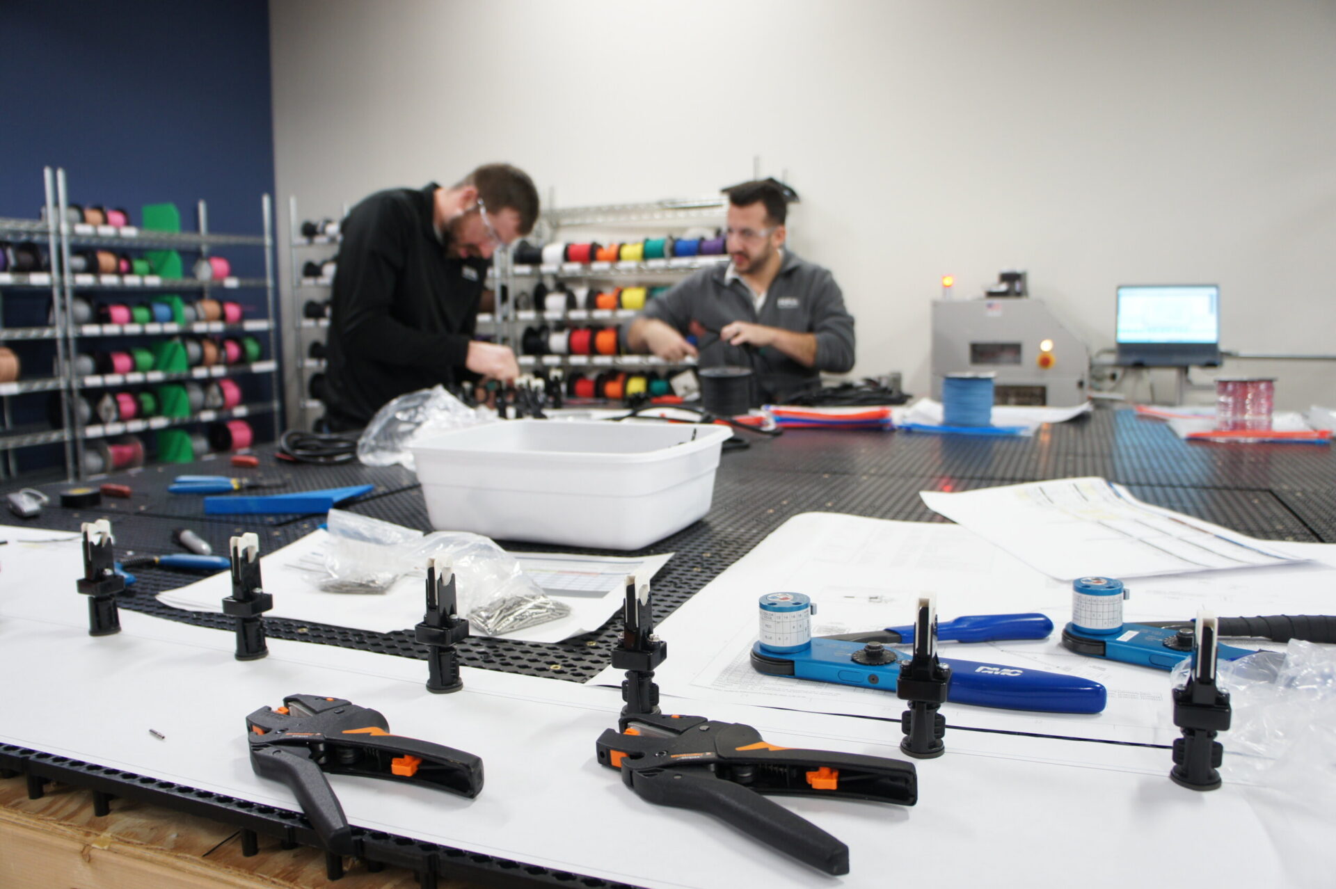 Two technicians working at a cluttered lab bench, assembling components with shelves of colorful spools in the background.