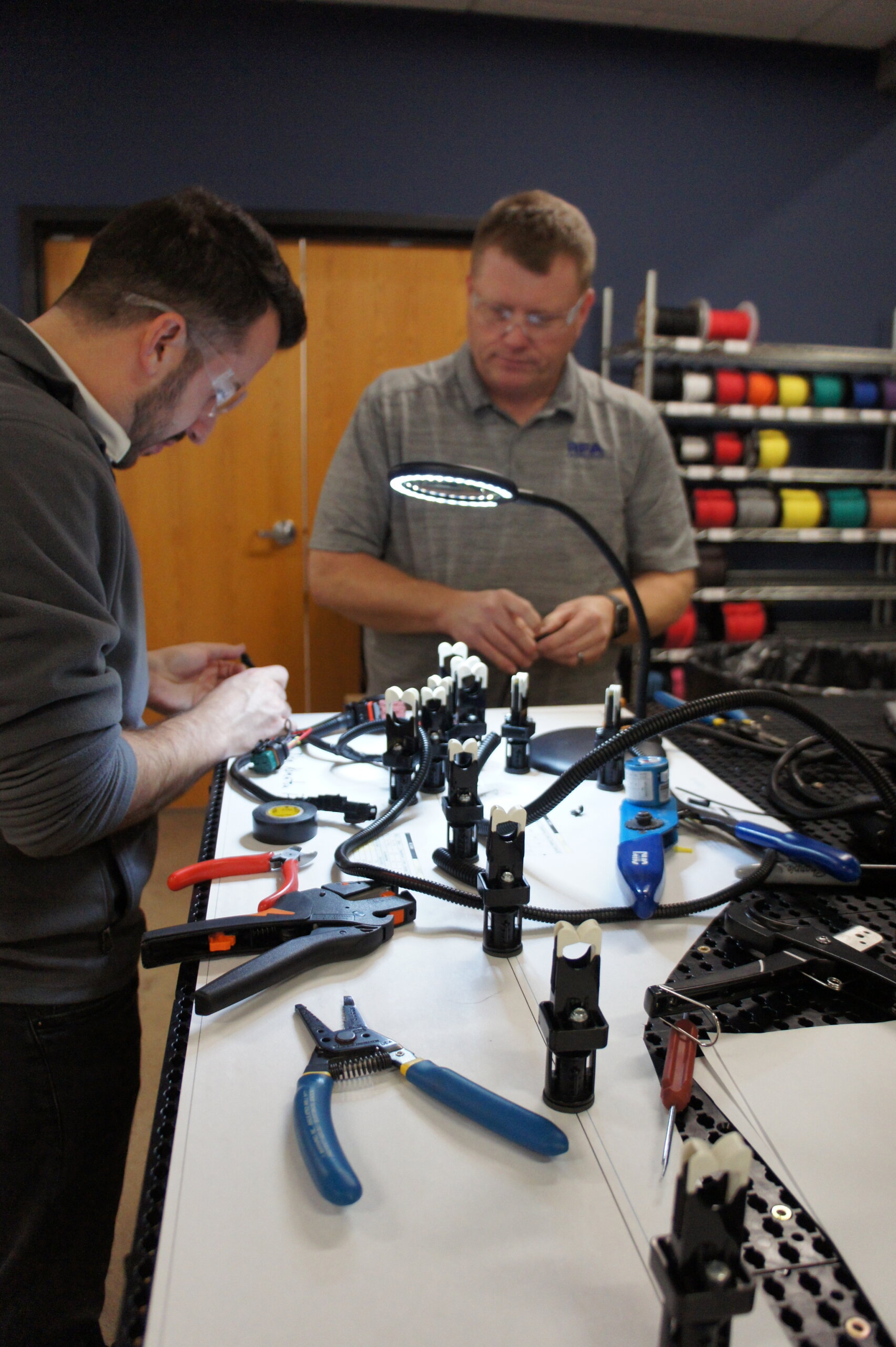 Two technicians assembling a prototype wire harness on a workbench with tools and components.