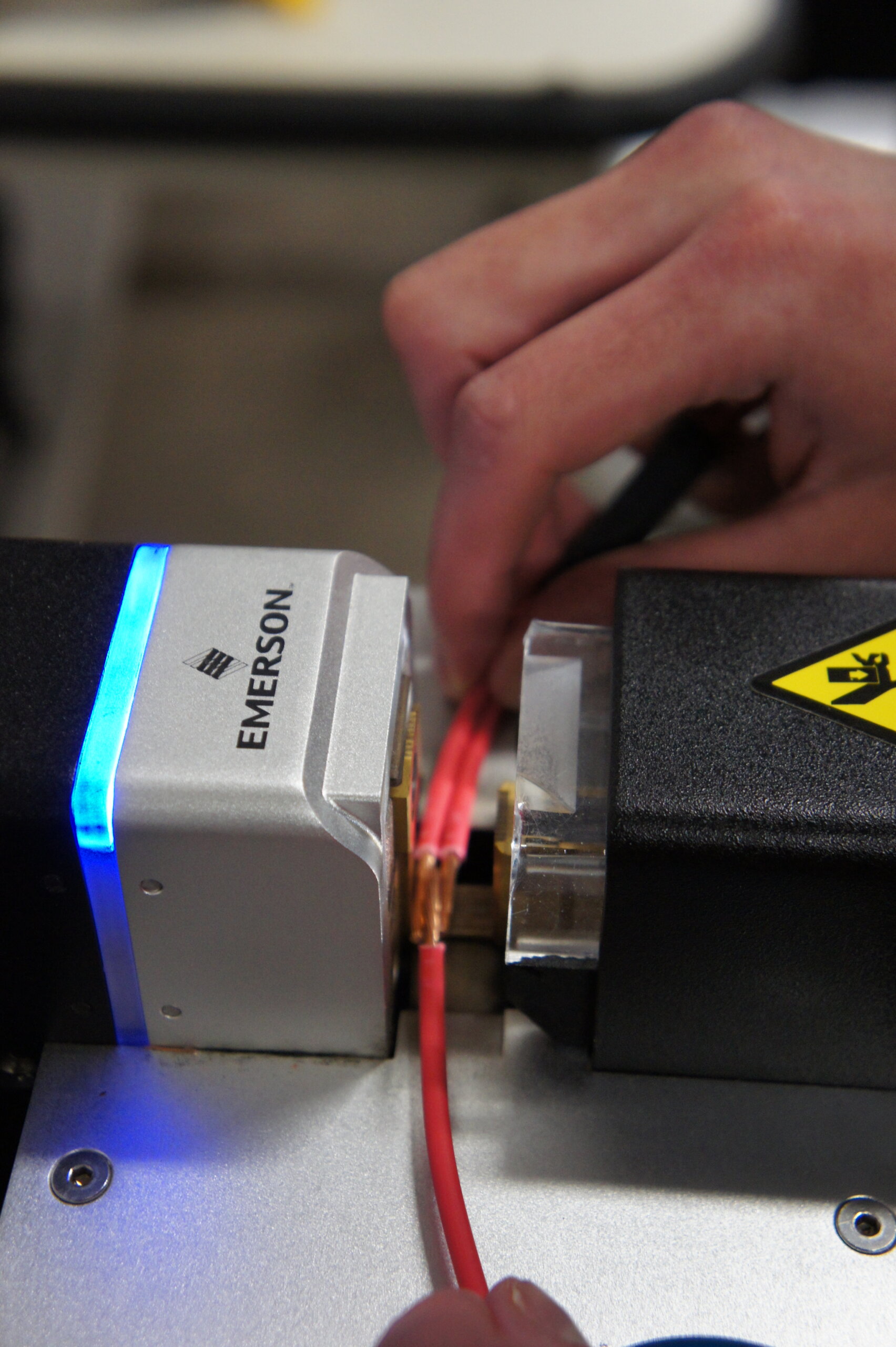 Close-up of a hand inserting a red wire into a metallic device with a blue LED glow, electronics wiring visible (Emerson branding).