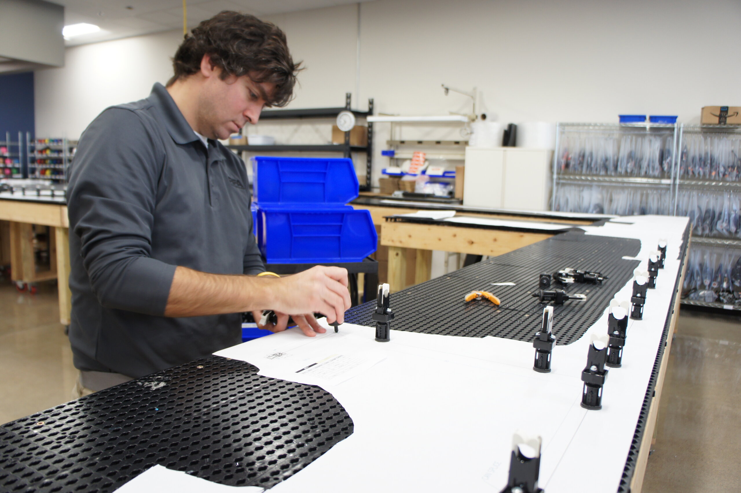 Man in a gray polo assembles small components at a perforated worktable in a workshop, with blue storage bins behind him.
