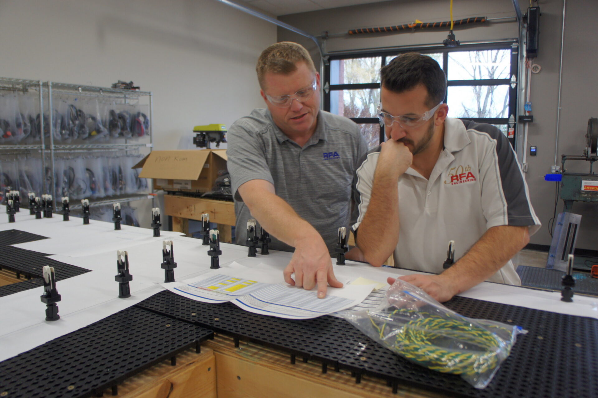 Two men in safety glasses lean over a long work table, reviewing papers amid a grid-like pegboard with small fixtures and parts nearby in a workshop.