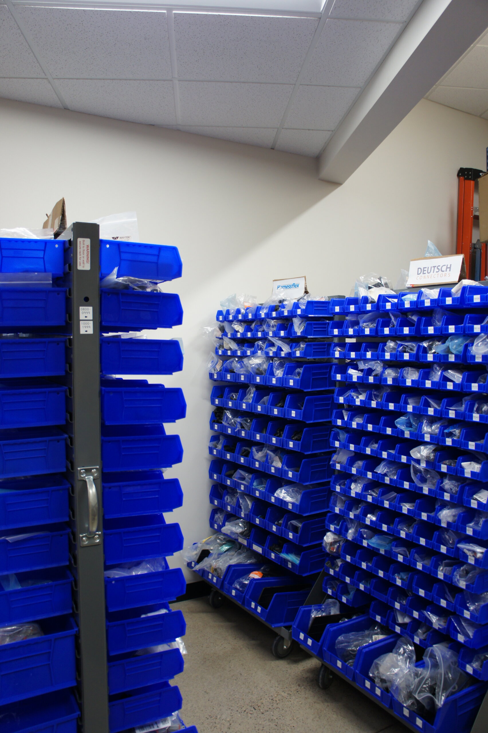 Storage room or workshop with tall metal racks filled with blue plastic bins and labeled compartments on both sides of a aisle.