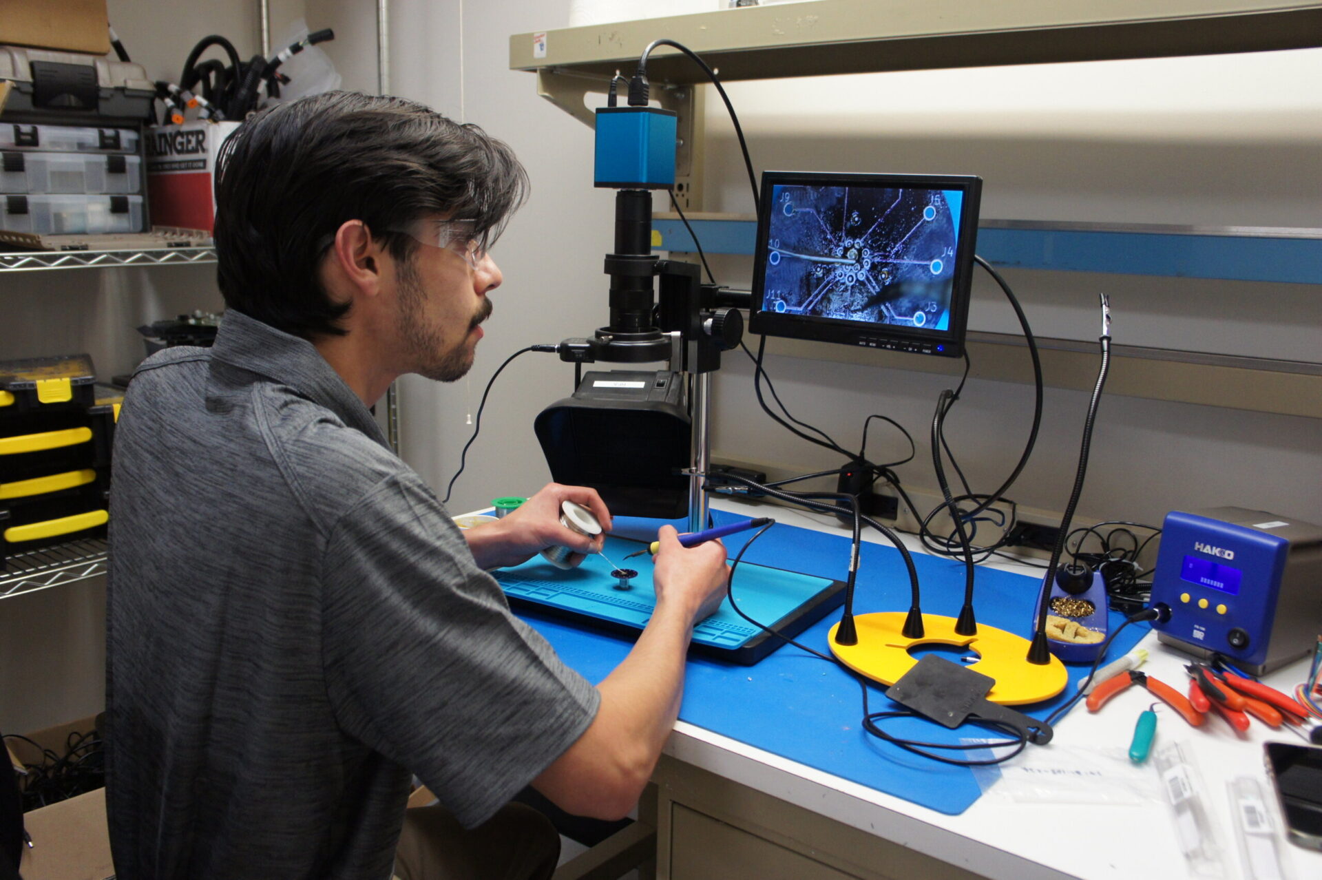 Man wearing safety glasses works at a blue lab bench, using tools while a monitor shows a magnified circular sample edge-to-edge image.