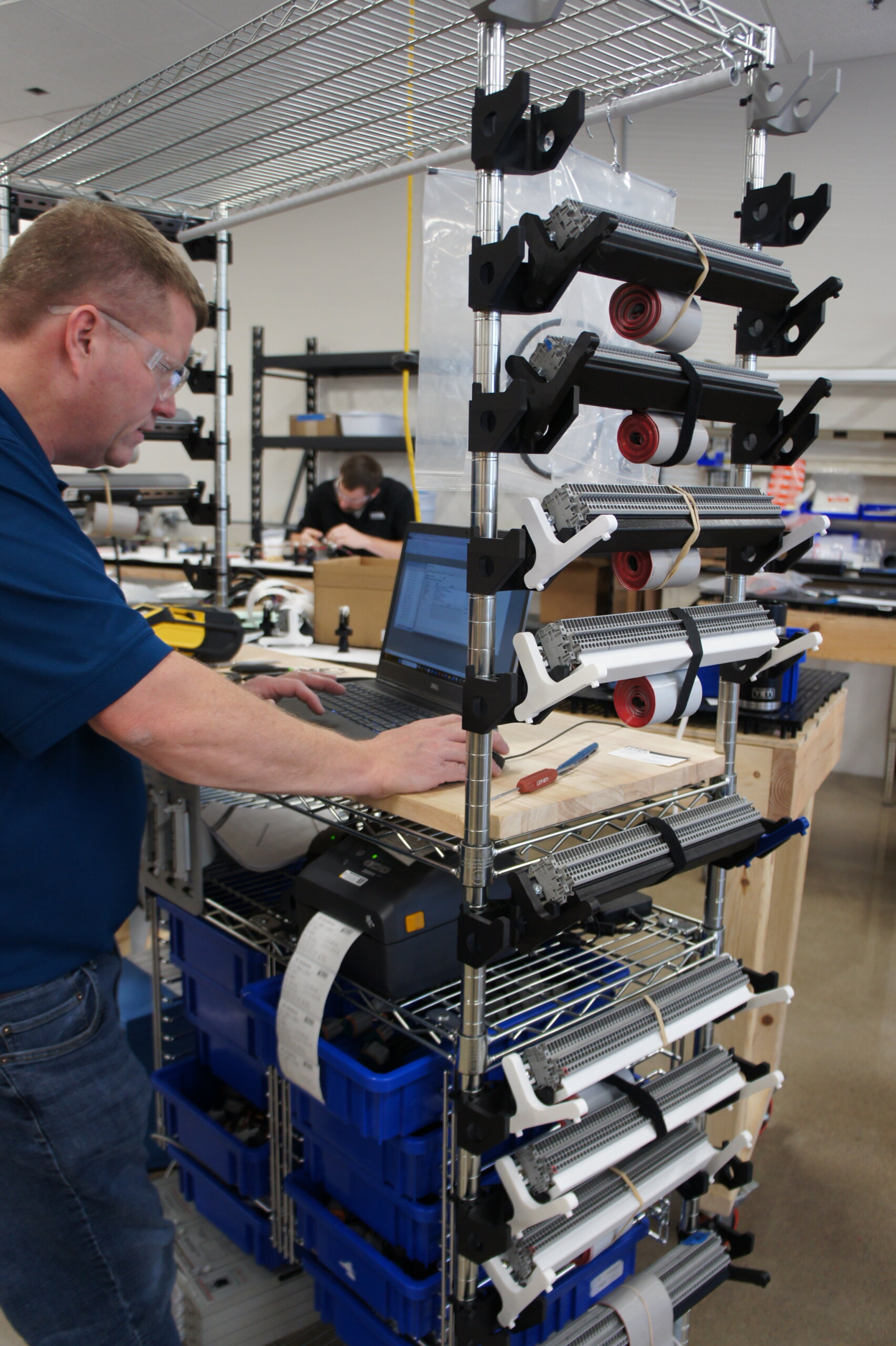 Man in safety glasses typing on a laptop beside a tall rack of cable harnesses and spools in a workshop.