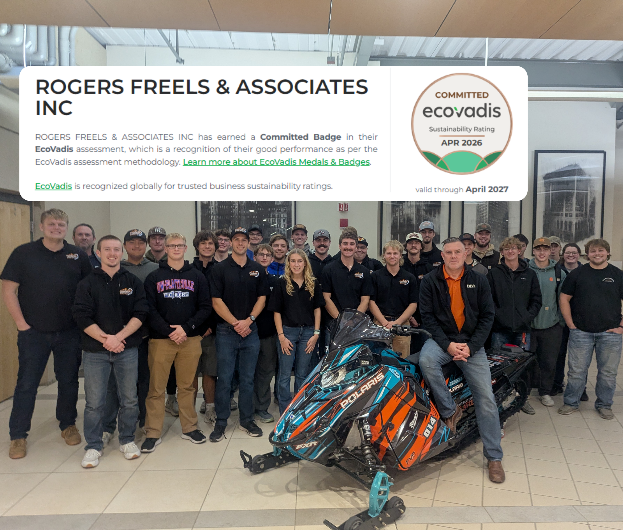 Group of people in matching dark shirts posing in a showroom with a blue‑orange Polaris snowmobile in the foreground, facing the camera.